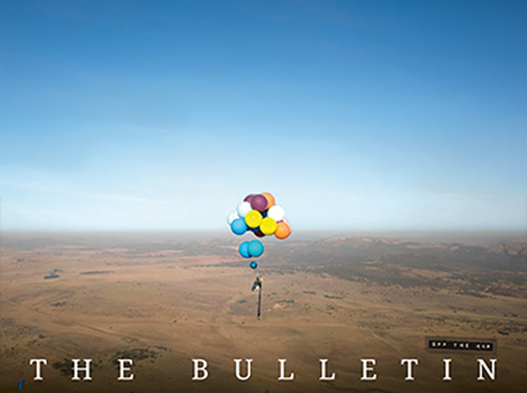 A person suspended beneath a cluster of colorful balloons over a vast open landscape.