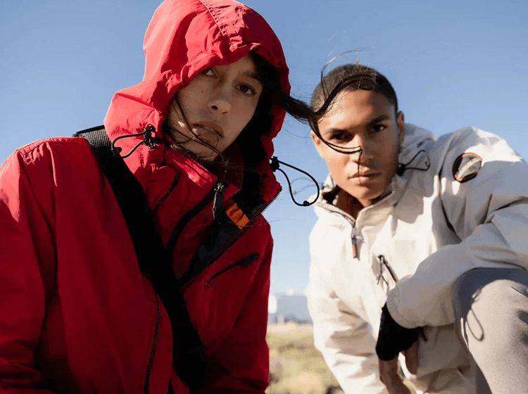 Two people wearing red and beige Napapijri jackets outdoors in windy seaside setting
