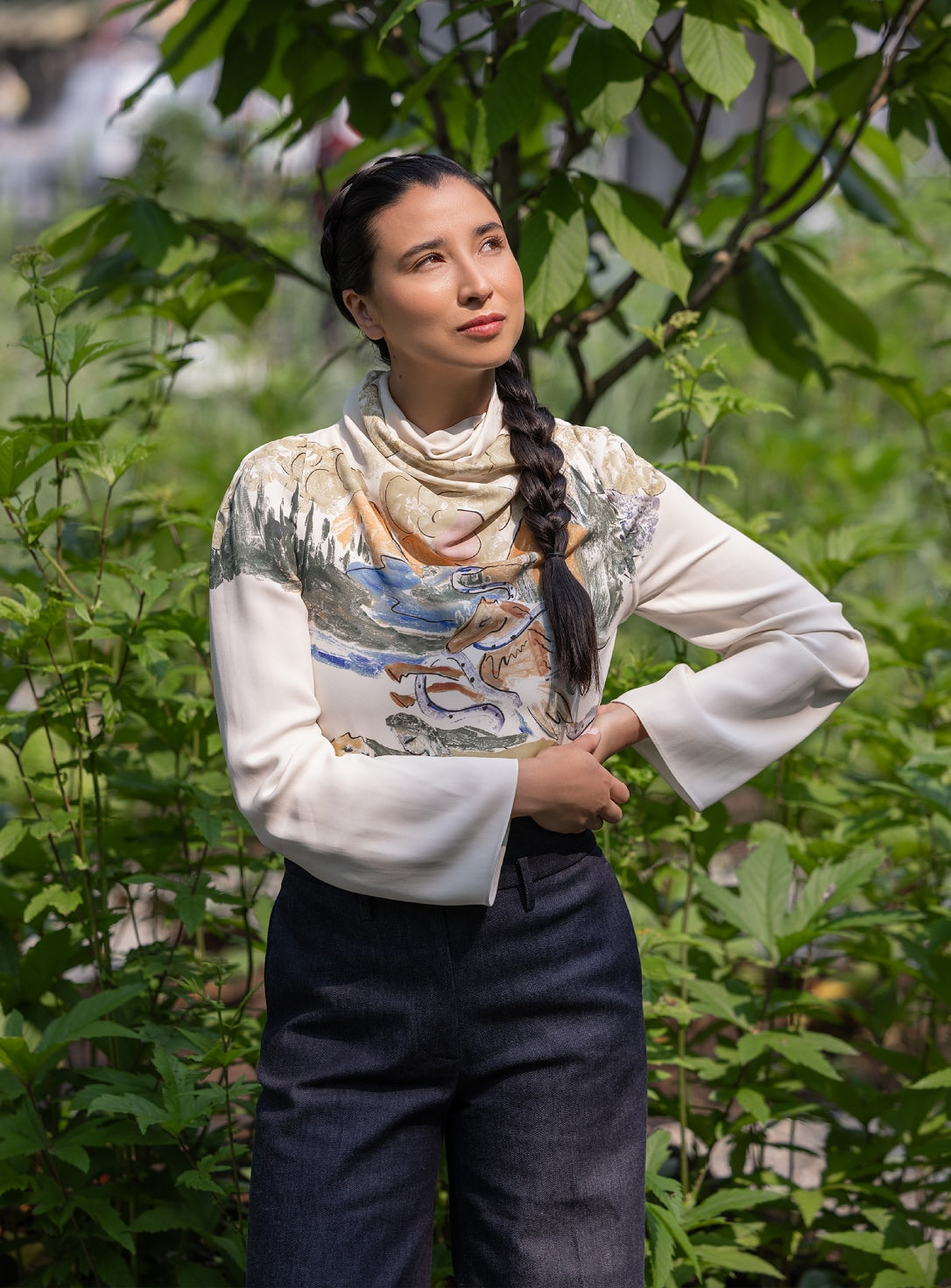 Portrait of a person standing in a green garden, wearing a printed long-sleeve top and scarf.