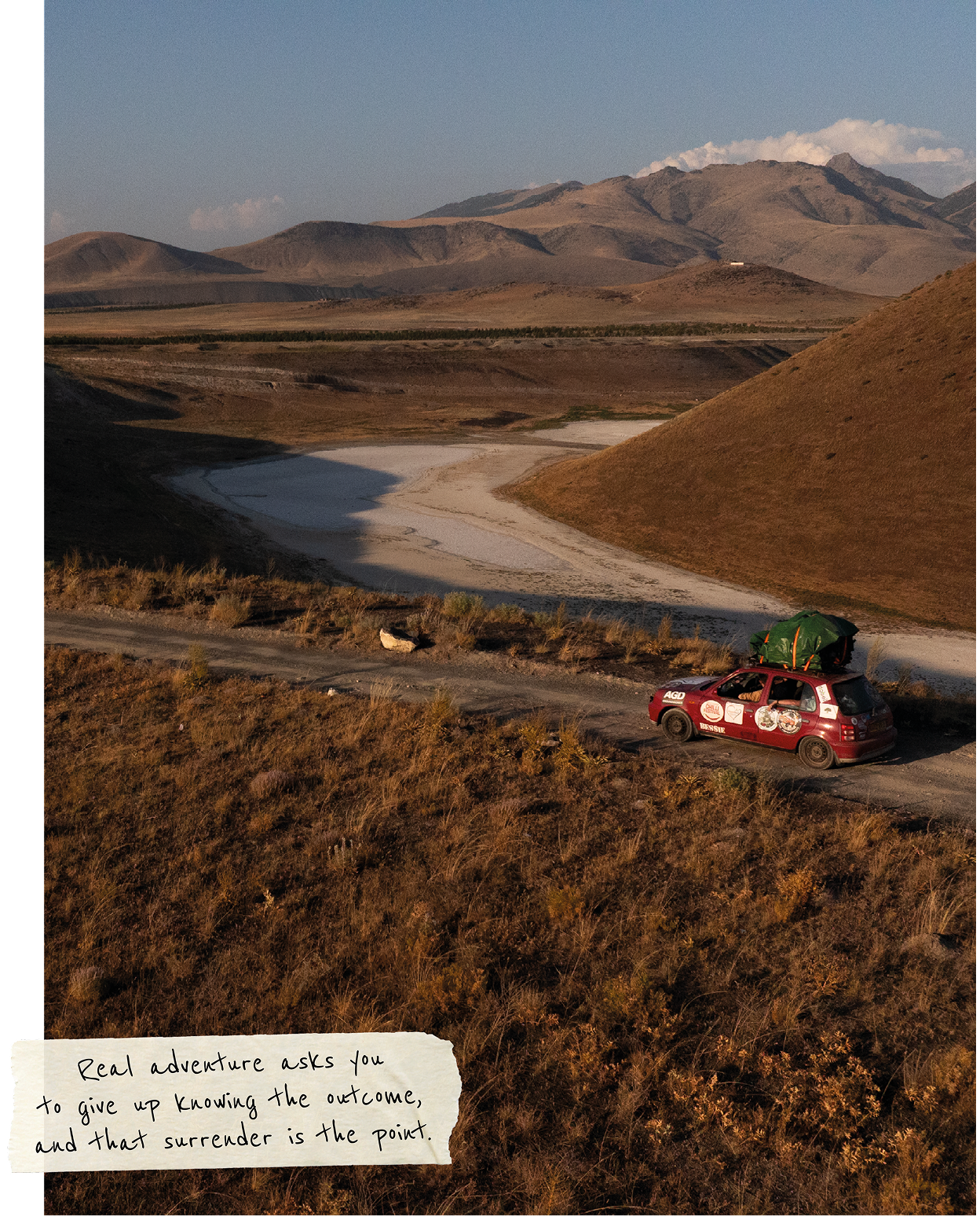 A rally car with roof cargo driving through a dry valley surrounded by hills.