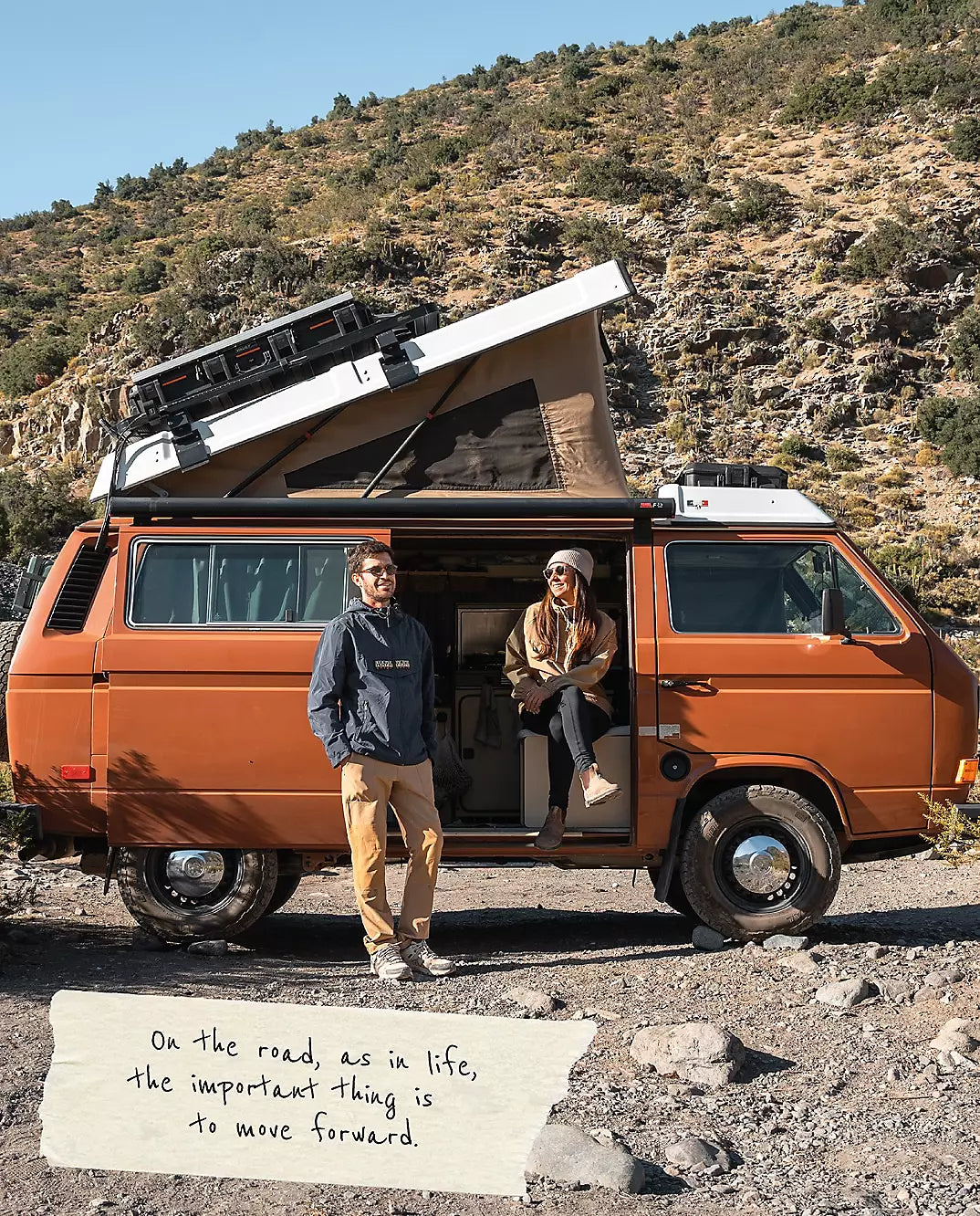 Two people standing beside camper van with roof tent in rocky terrain