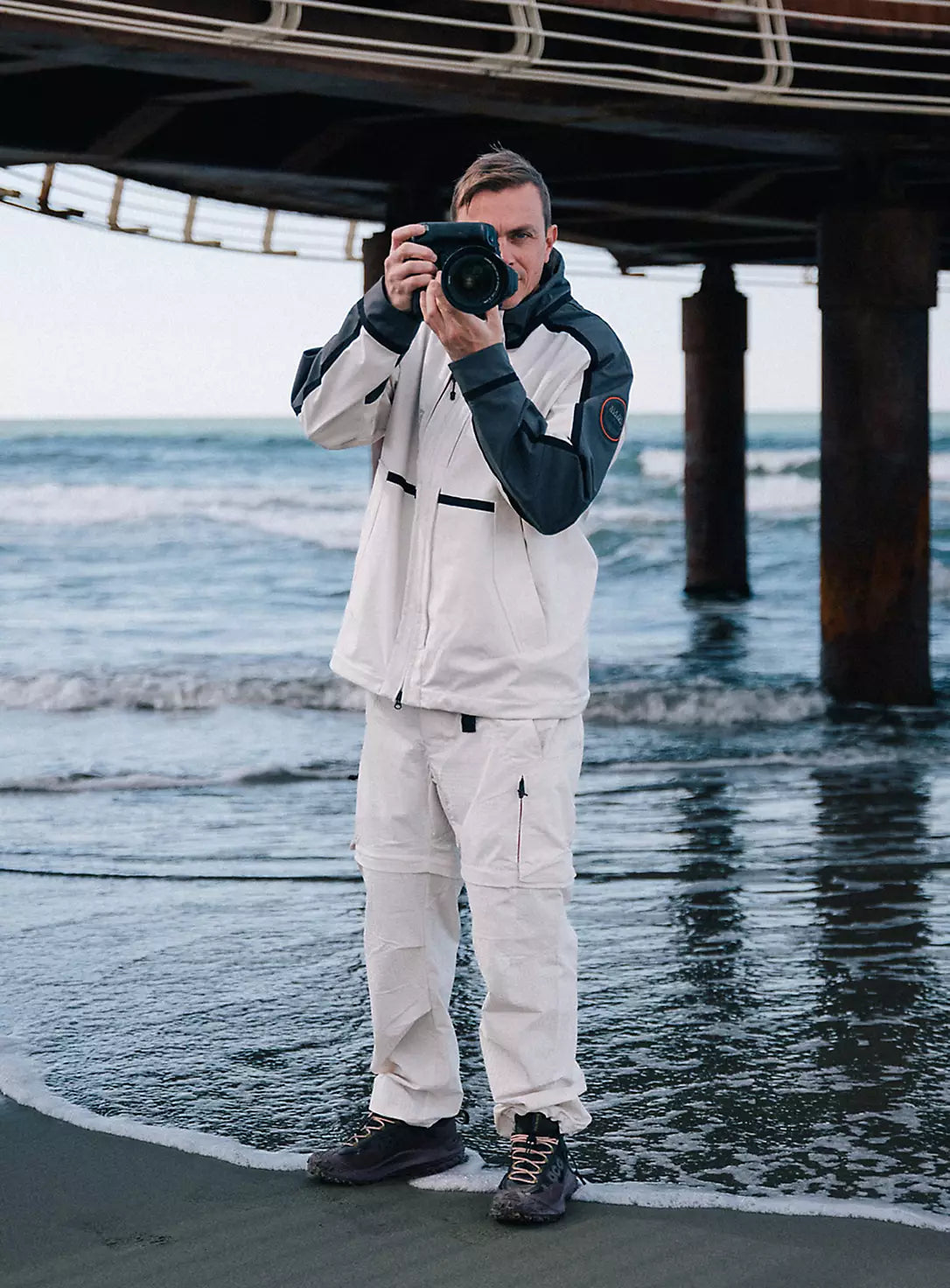 A person wearing Napapijri technical jacket while photographing shoreline beneath wooden pier